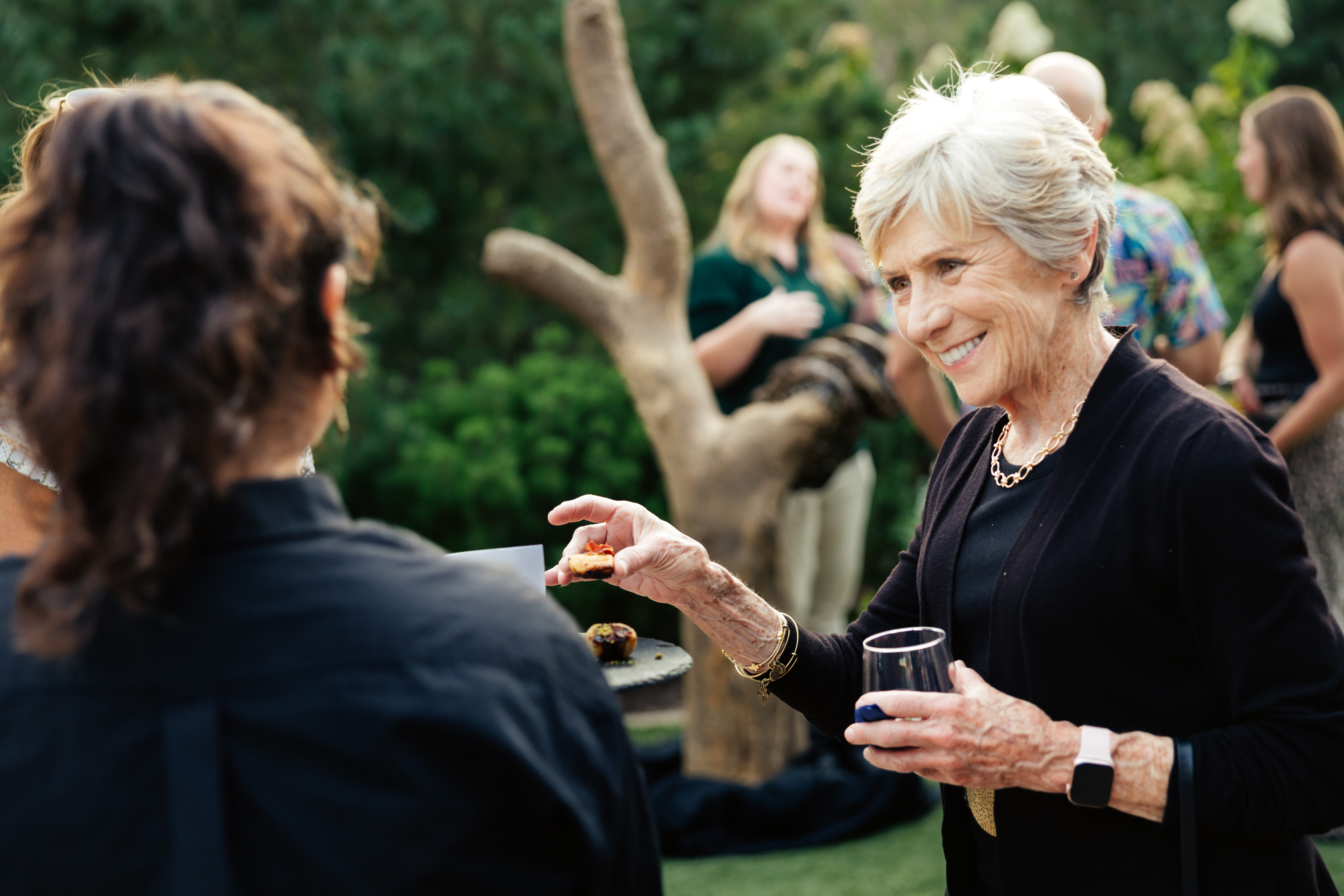 An older woman smiles and reaches for food at an outdoor gathering, holding a drink in her other hand.
