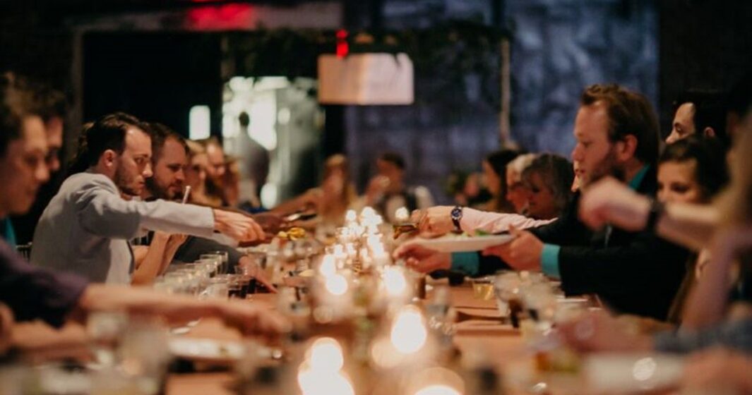 People seated at a long, candlelit table share food and drinks in a dim, festive atmosphere.