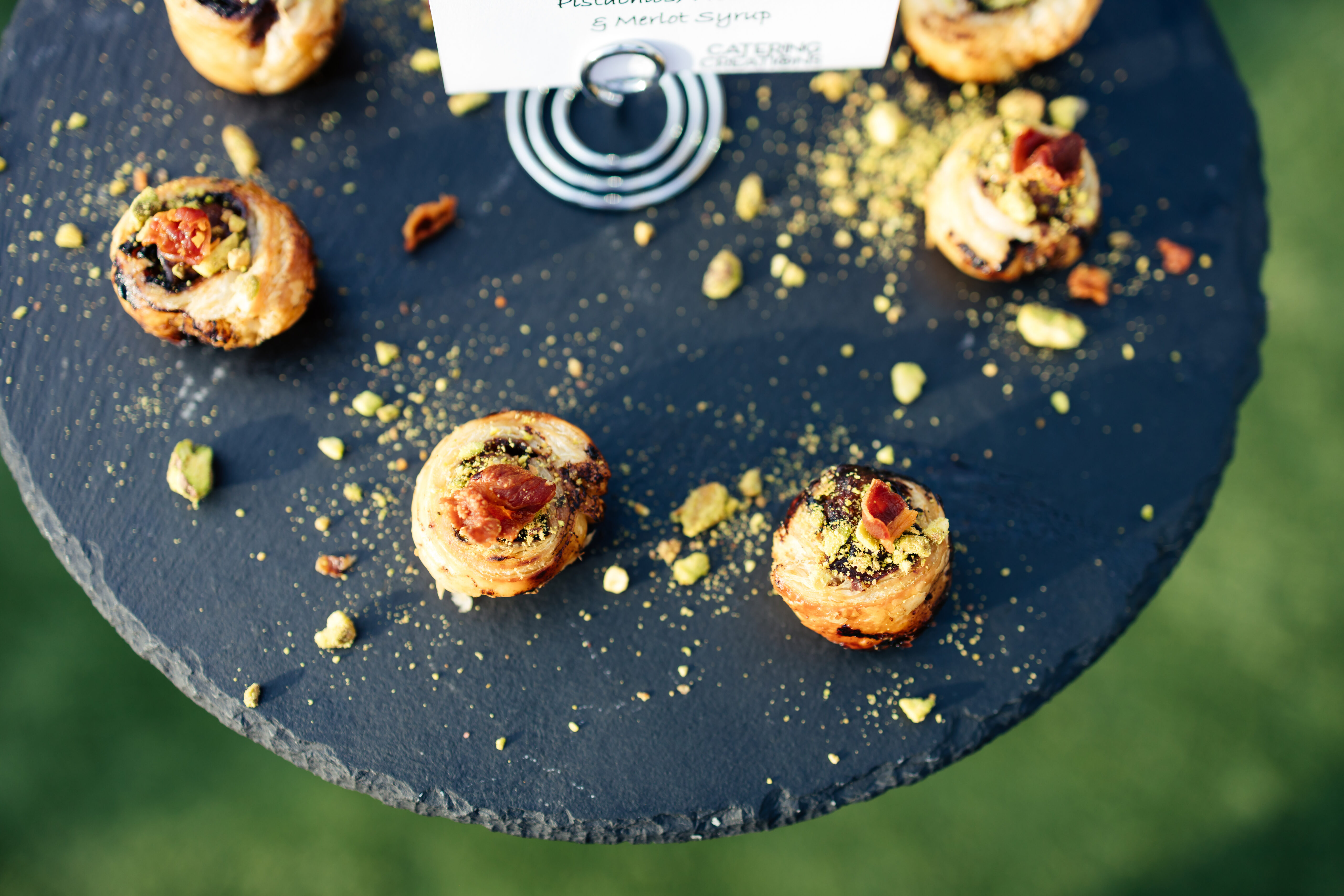 Mini pastries topped with chopped pistachios and fruit, arranged on a round black slate serving tray.