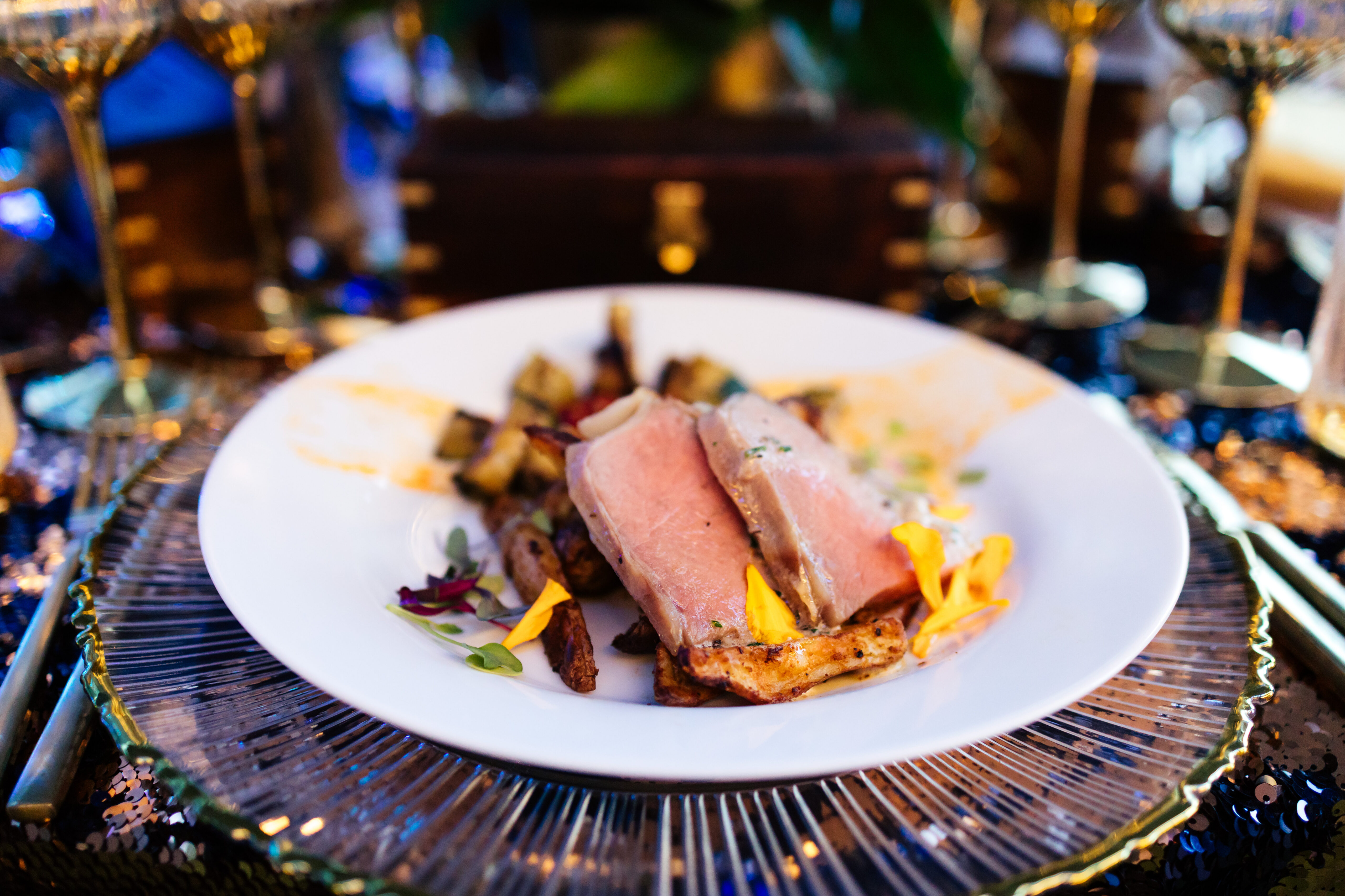 Sliced meat on roasted vegetables, garnished with herbs and edible flowers, served on a white plate with gold accents.
